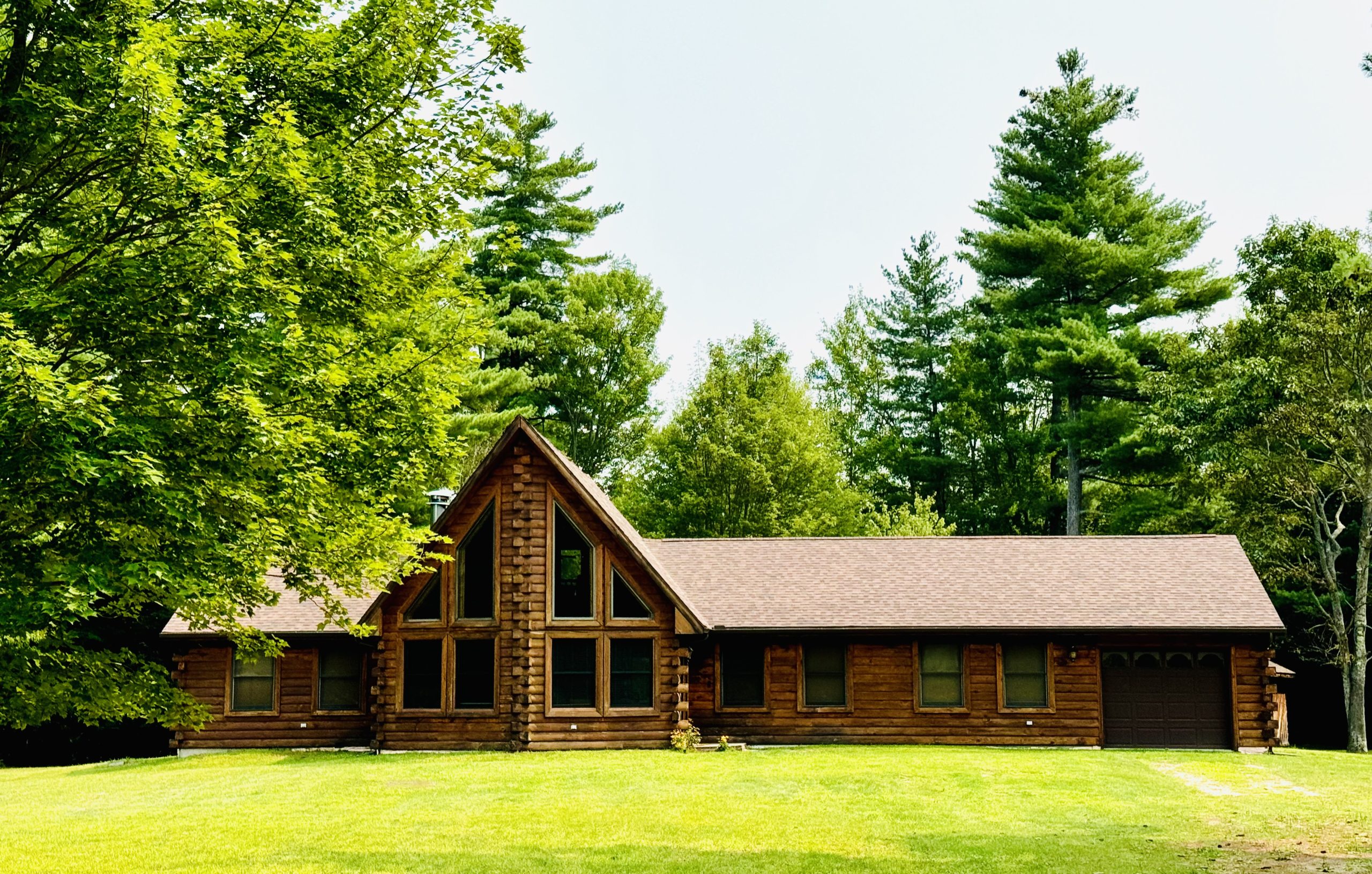 Rustic log cabin home with large A-frame windows, nestled in a lush green forest with manicured lawn and pine trees, perfect for vacation rental in the Adirondacks or Lake Placid region