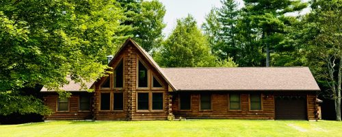 Rustic log cabin home with large A-frame windows, nestled in a lush green forest with manicured lawn and pine trees, perfect for vacation rental in the Adirondacks or Lake Placid region